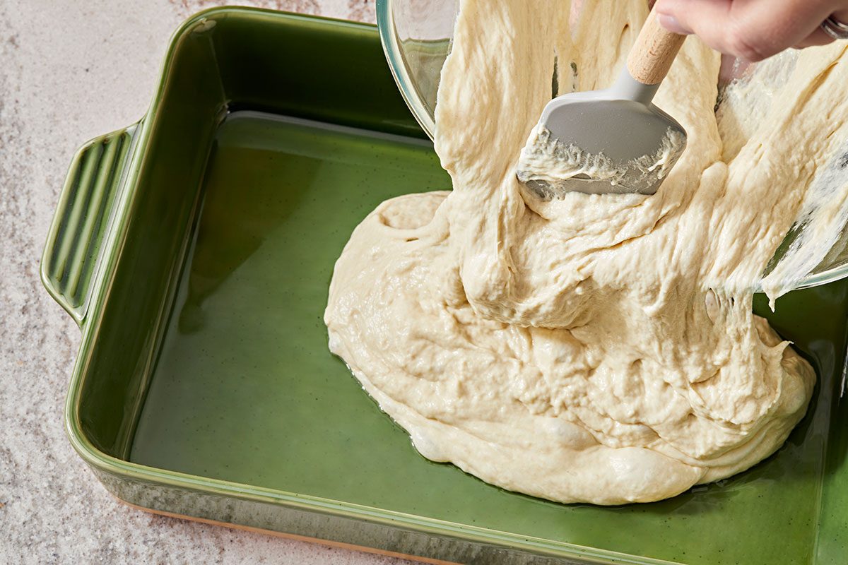 High-angle shot of a person using a spatula to spread thick; creamy batter from a bowl into a greased green rectangular baking dish.