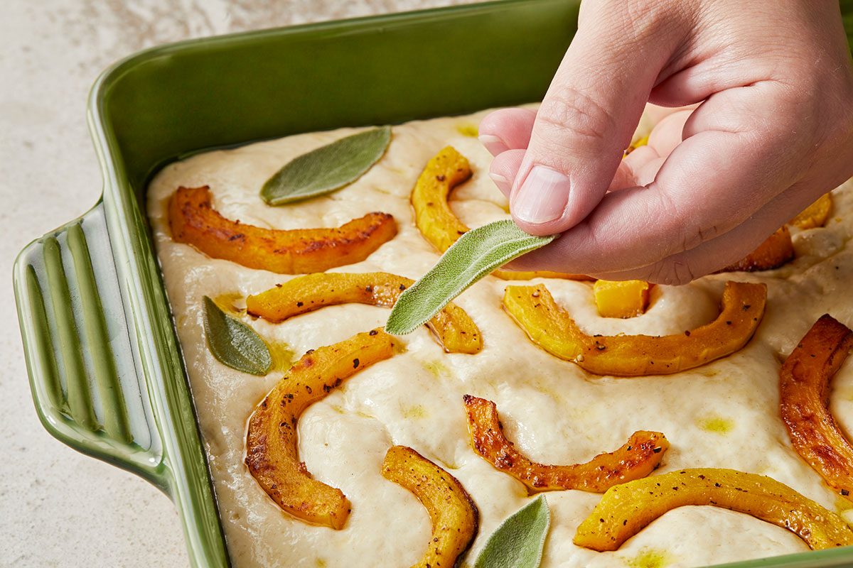 Close-up shot of a hand placing a fresh sage leaf onto unbaked focaccia dough topped with roasted squash slices in a green baking dish.