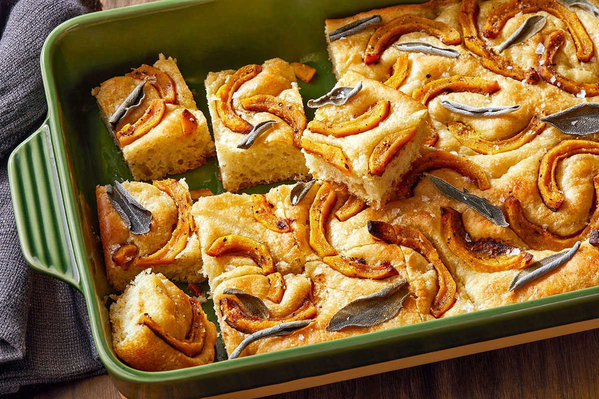 Close-up shot of Squash and Sage Focaccia in a green rectangular baking dish; cut into squares with a few pieces lifted to reveal the soft; airy texture inside.