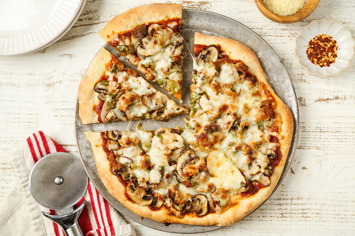 A homemade pizza topped with mushrooms, cheese, and green peppers sits on a round metal tray. Three slices are cut. A pizza cutter, a red striped napkin, and small bowls of cheese and chili flakes are nearby.
