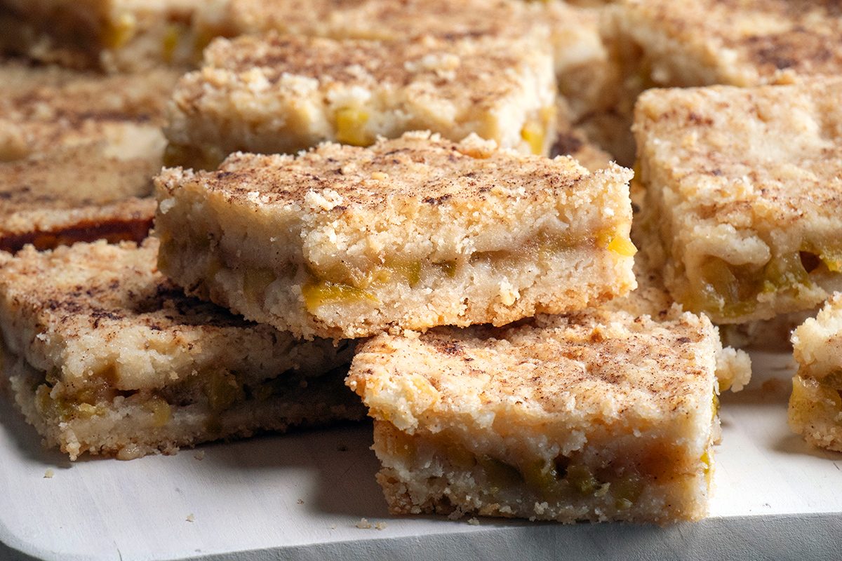 A close-up of crumbly dessert bars stacked on a plate, featuring a golden-brown crust and visible filling, likely made with fruit or a similar sweet filling.