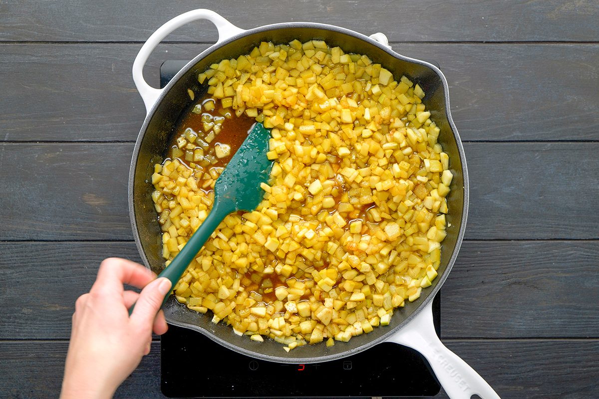 A hand stirs a skillet filled with diced apples cooking in a brown sauce, using a green spatula. The pan is on a stovetop over a dark wooden surface.