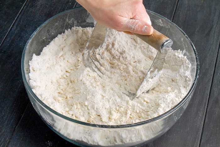 A hand uses a pastry blender to mix flour in a glass bowl on a dark wooden surface.