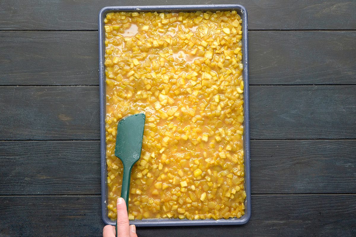 A hand uses a green spatula to spread a chunky yellow mixture evenly in a rectangular baking tray on a dark wooden surface.