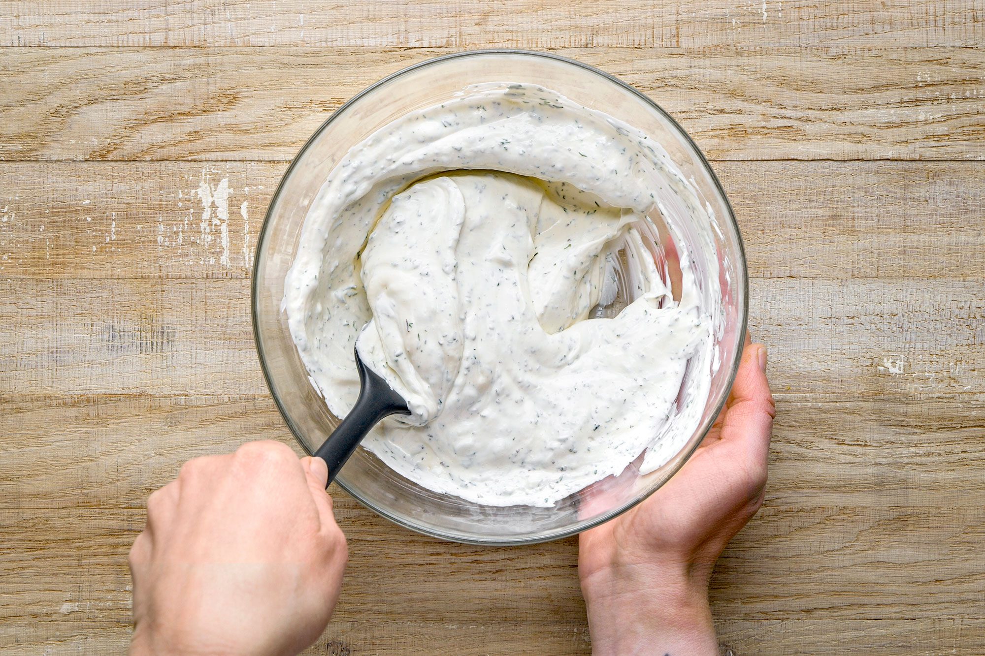 Overhead shot of a person stirring creamy white sauce with herbs in a glass bowl using a black spatula on a wooden surface;