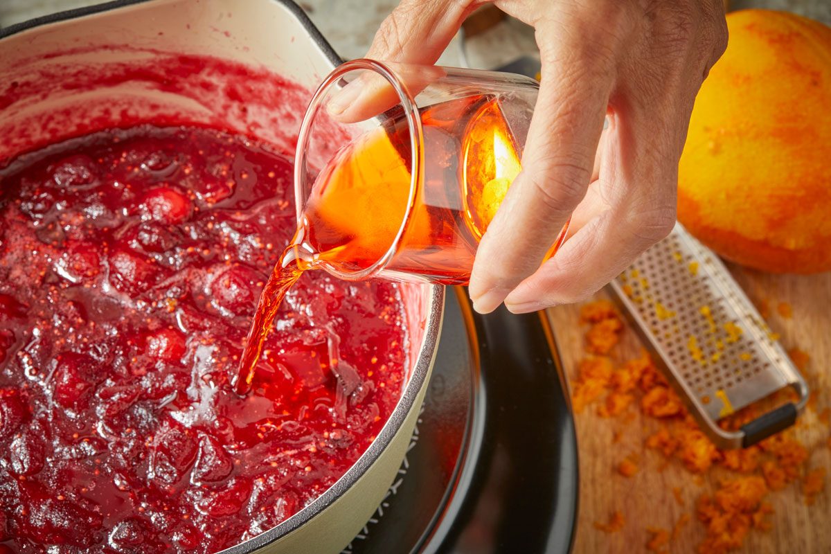 Close-up of a hand pouring Aperol from a glass into a saucepan of simmering cranberry sauce, with an orange, zester, and scattered orange zest on the wooden surface nearby;