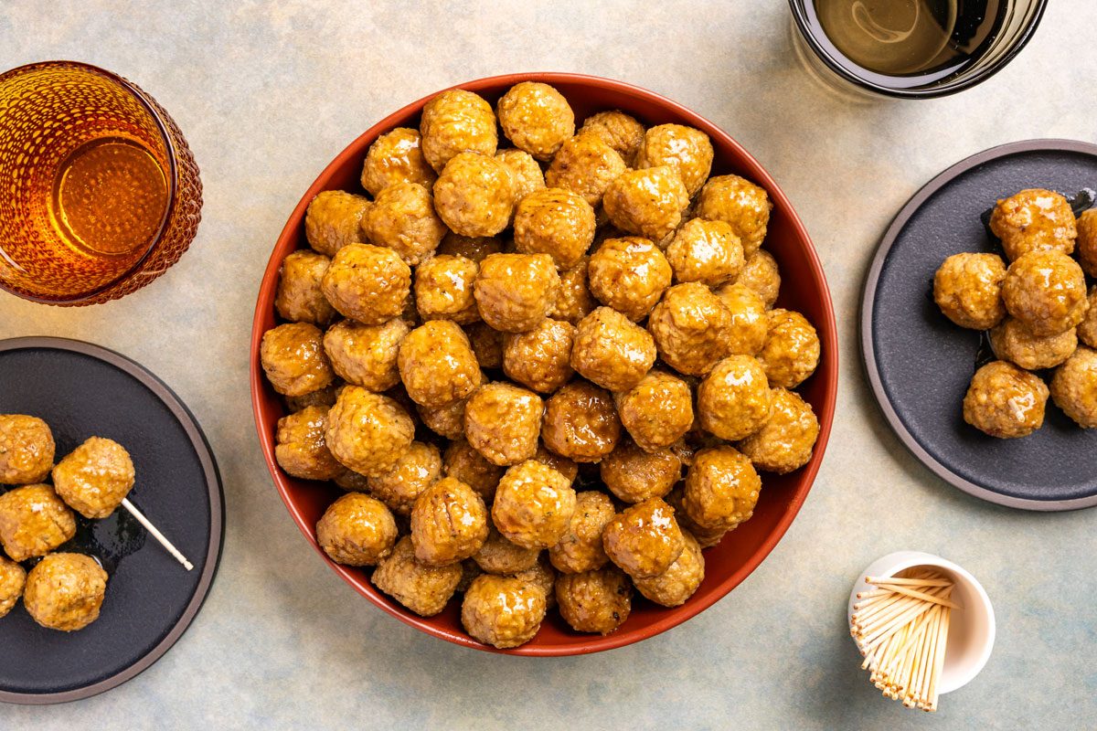 This is an overhead shot; a big red bowl of glazed meatballs sits on a table; small plates; drinks; toothpicks; wooden surface