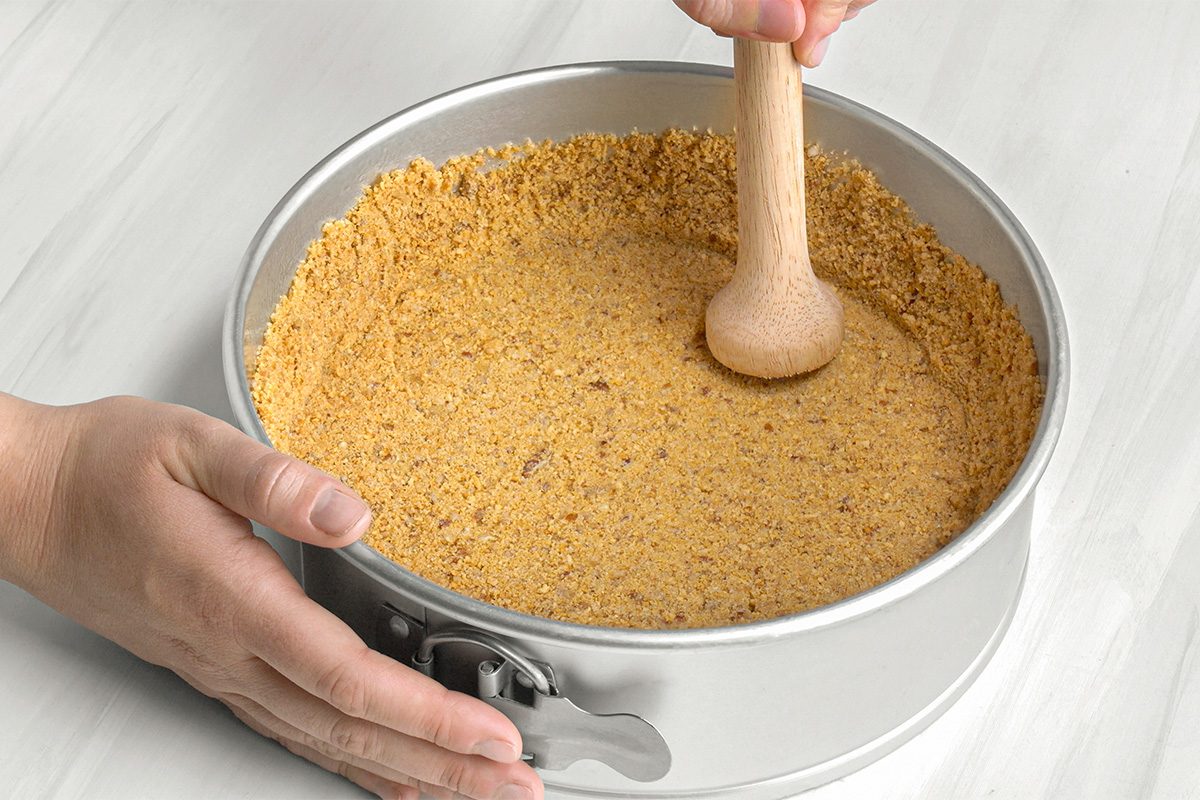A person presses a graham cracker crust into a round springform pan using a wooden tamper, preparing a dessert base on a light-colored surface.