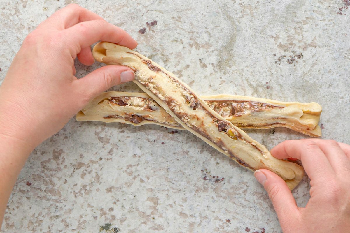 Two hands twisting a strip of dough with visible nuts and chocolate pieces on a light, speckled surface. Another similar dough strip lies underneath.
