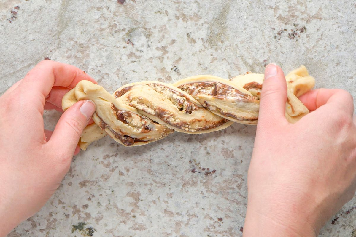 Two hands are holding and twisting a strip of dough with a visible chocolate or nut filling, forming a braided pattern on a light-colored countertop.