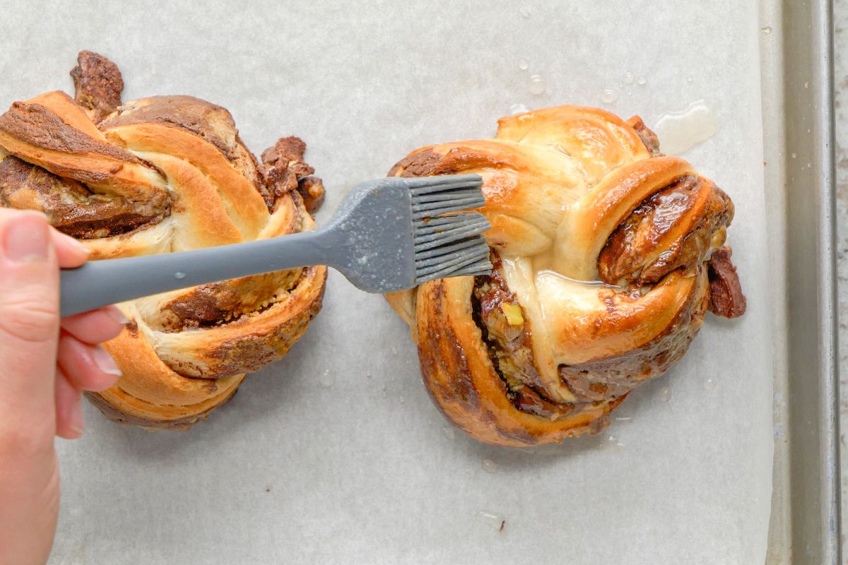 A hand uses a pastry brush to glaze two twisted, round babka buns filled with chocolate and nuts on a parchment-lined baking sheet.