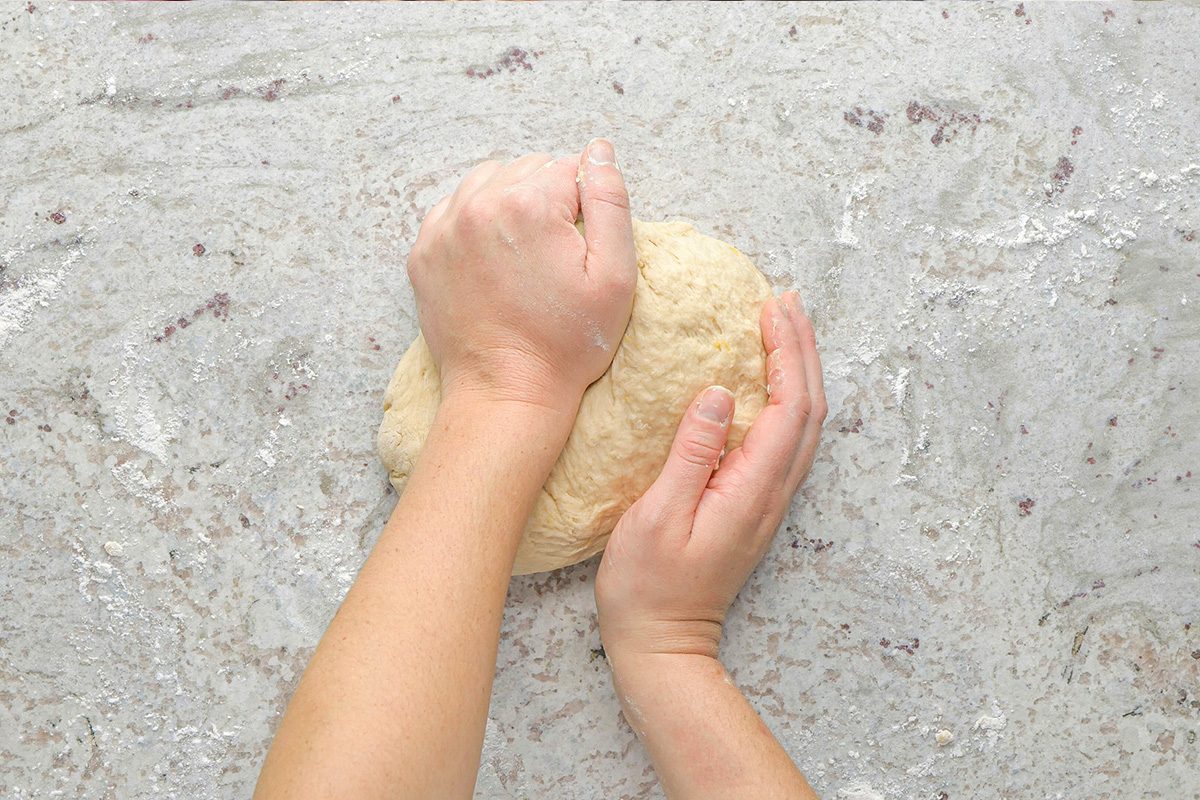 A pair of hands kneads a ball of dough on a lightly floured, gray countertop.