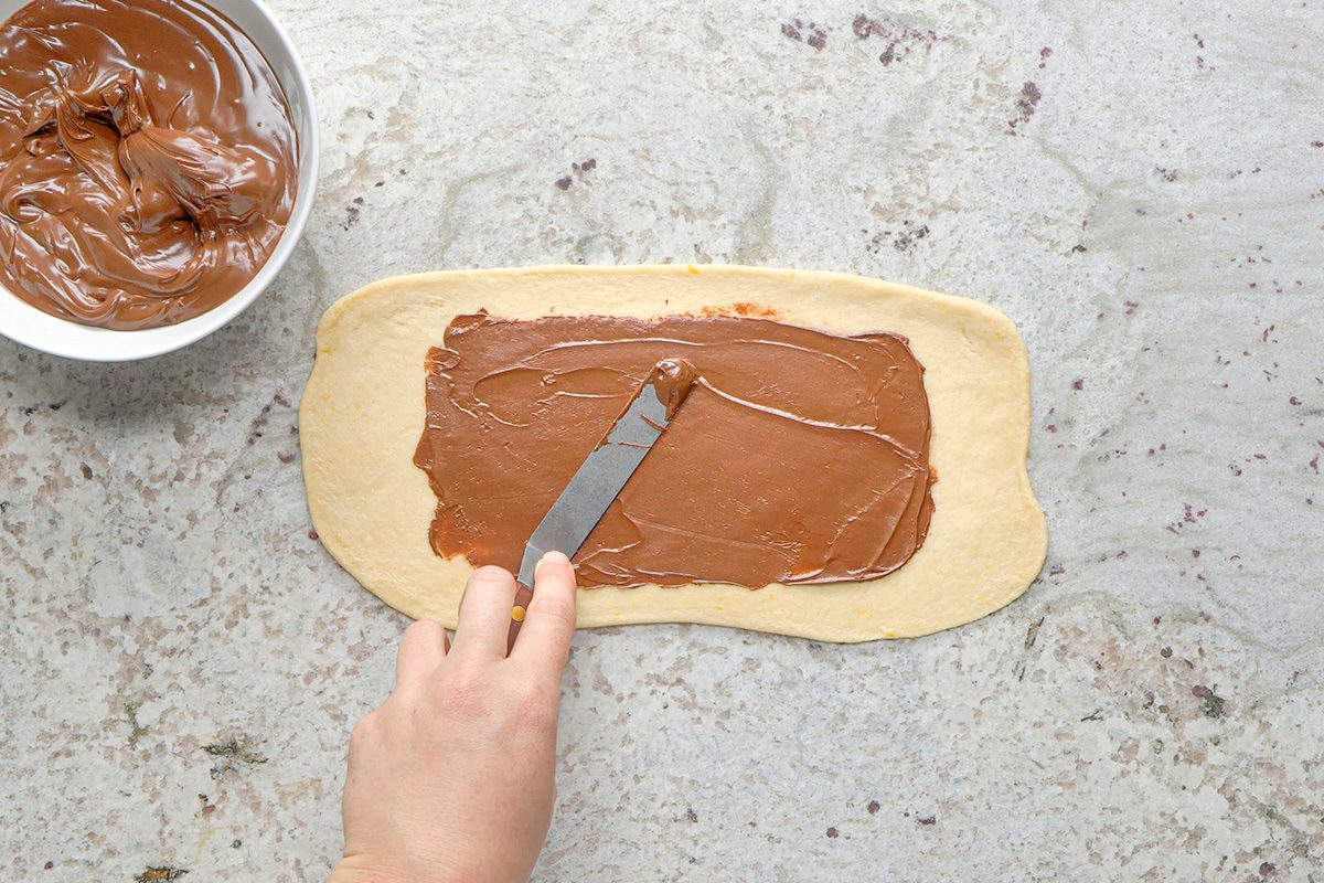 A hand spreads chocolate hazelnut spread onto a rectangular piece of dough with a metal spatula; a bowl of more spread is nearby on a gray countertop.