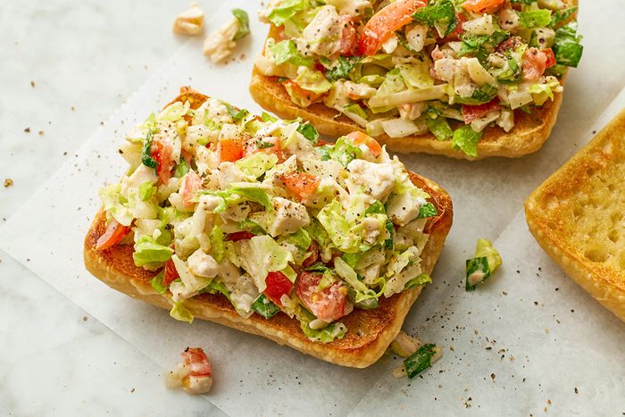 This is a top-down shot; two open-faced toasted sandwiches are topped with creamy chicken salad, chopped lettuce; tomatoes; and herbs, placed on parchment paper over a white marble background