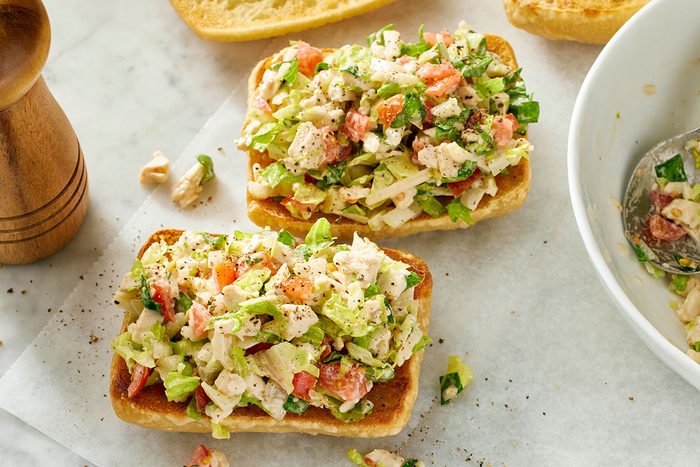 vThis is an overhead shot showing two open-faced chicken salad sandwiches on toasted bread atop parchment; nearby are a bowl of extra salad with a spoon and a pepper grinder on the left