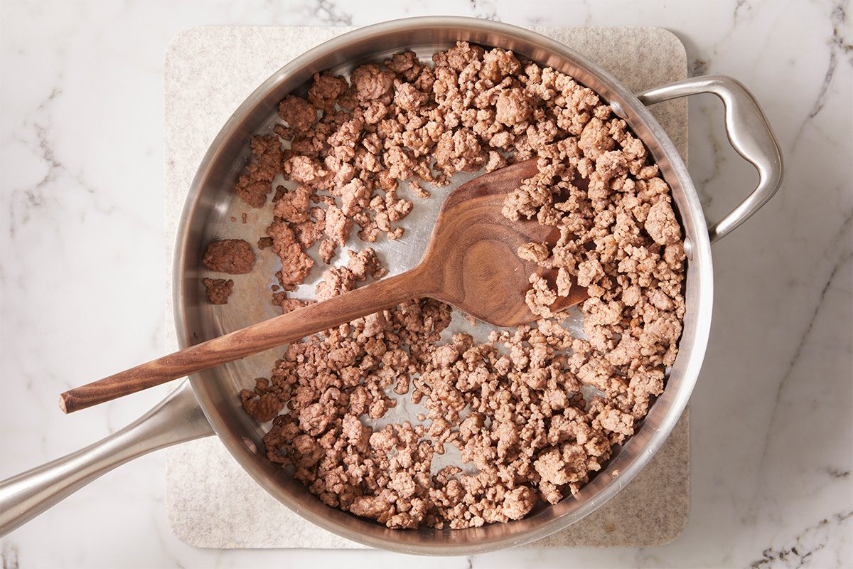 A stainless steel skillet with cooked, crumbled ground beef and a wooden spoon resting inside, set on a light-colored countertop.