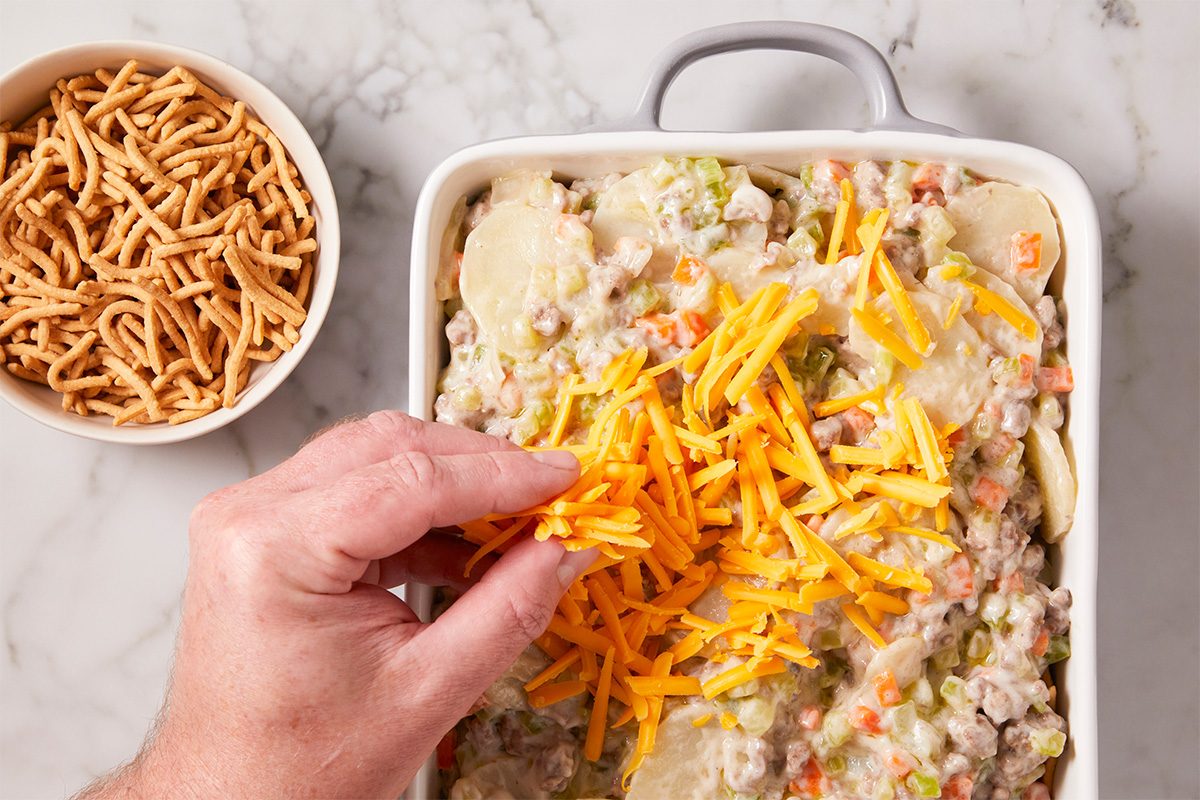 A hand sprinkles shredded cheddar cheese over a casserole filled with creamy vegetables and ground meat. Next to the dish is a bowl of crunchy chow mein noodles on a marble countertop.