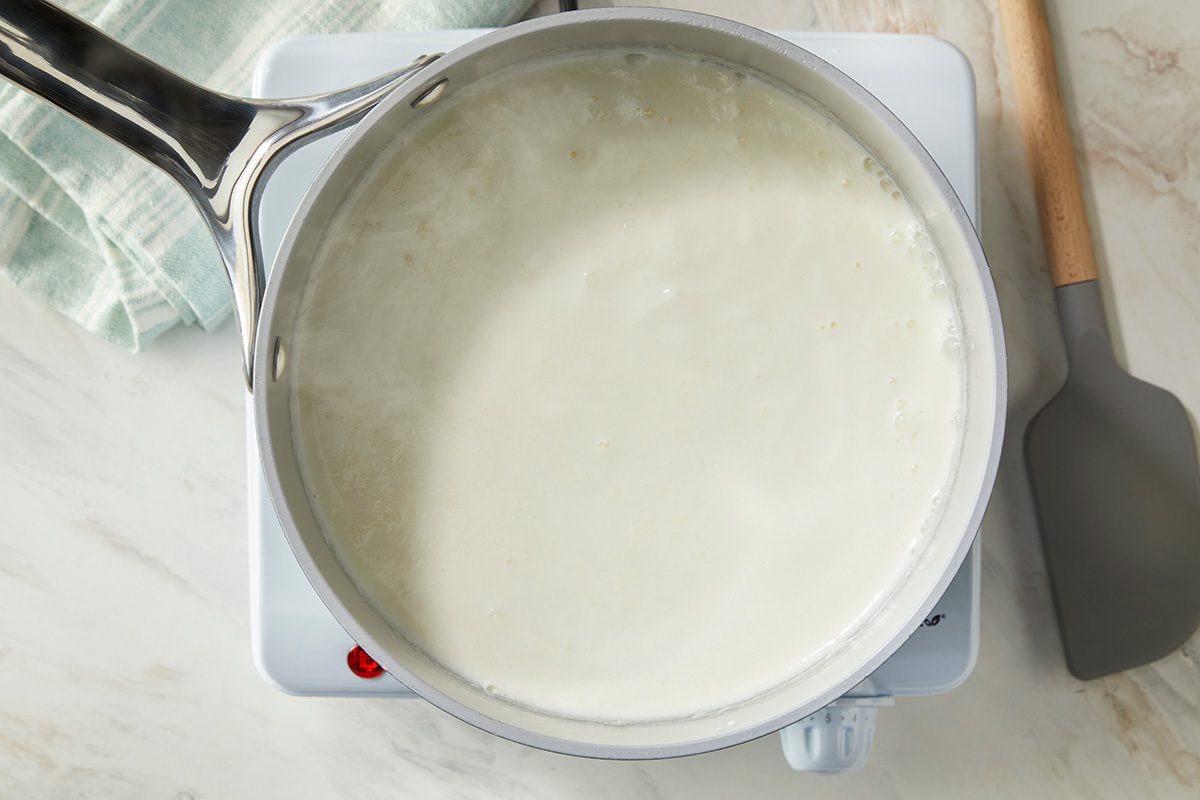 A saucepan filled with milk is heating on a stovetop. A gray spatula lies next to the pot on a light-colored countertop, and a light green striped kitchen towel is partially visible in the corner.
