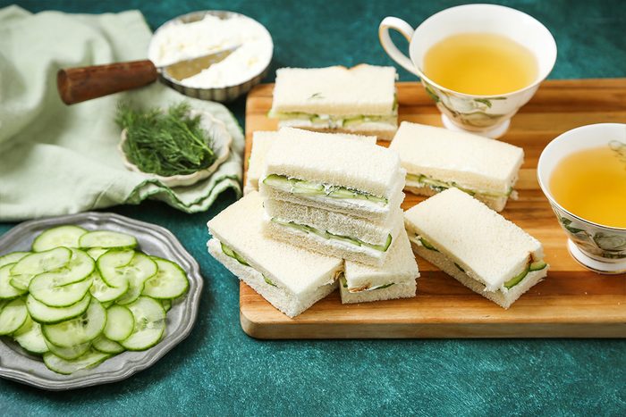 A wooden tray with stacked cucumber tea sandwiches sits next to two cups of tea. Nearby are a plate of sliced cucumbers, a bowl of spread, and fresh dill on a green tablecloth.