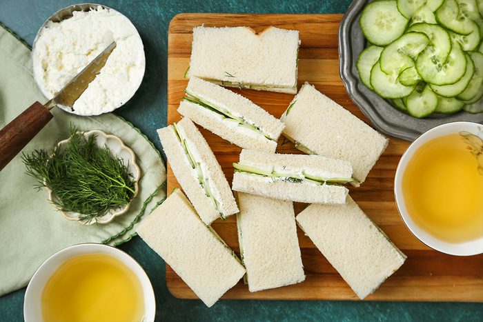 Rectangular cucumber tea sandwiches on a wooden board, surrounded by bowls of cucumber slices, herbs, a dish of soft cheese, and two cups of tea on a green table.