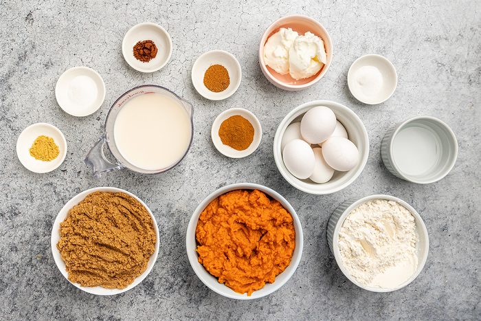 Various baking ingredients in bowls on a gray surface, including brown sugar, pumpkin puree, flour, eggs, milk, spices, baking powder, baking soda, shortening, and water, all arranged neatly for a recipe.