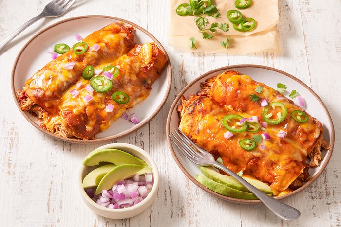 Two plates of cheesy enchiladas topped with sliced jalapeños, chopped red onions, and green onions sit on a white wooden table. A small bowl of avocado slices and red onion is nearby, along with extra garnishes.