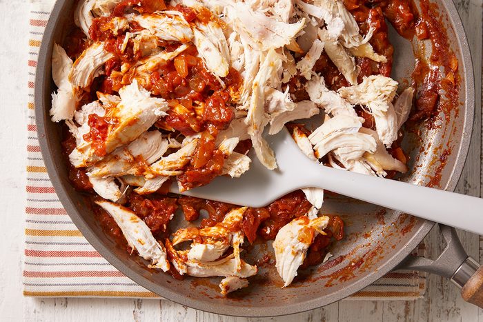 Shredded chicken mixed with tomato sauce in a skillet, with a white spatula resting inside. The pan sits on a striped cloth placed on a light wooden surface.