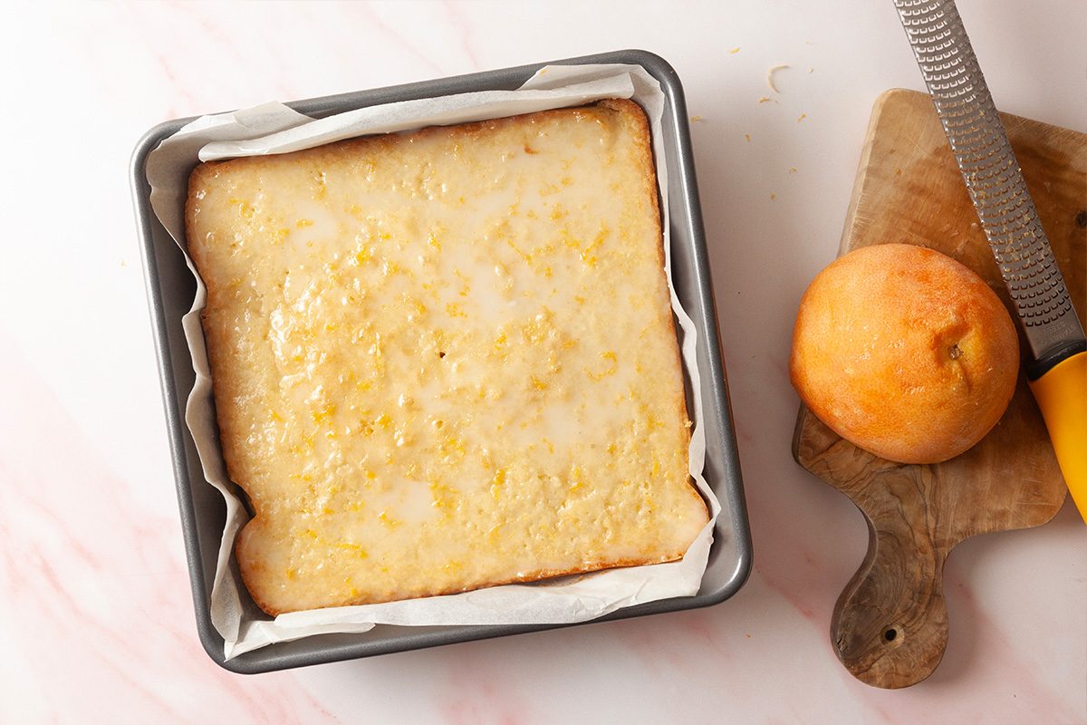 A square lemon bar topped with glaze sits in a parchment-lined baking pan. Beside it, a zested lemon and a grater rest on a small wooden cutting board.
