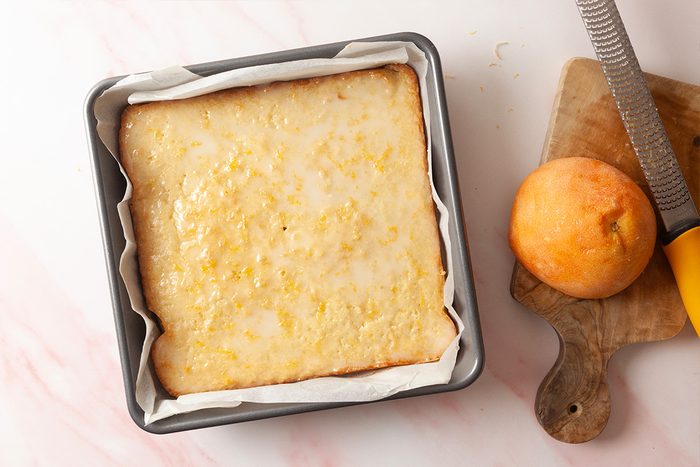 A square lemon bar topped with glaze sits in a parchment-lined baking pan. Beside it, a zested lemon and a grater rest on a small wooden cutting board.