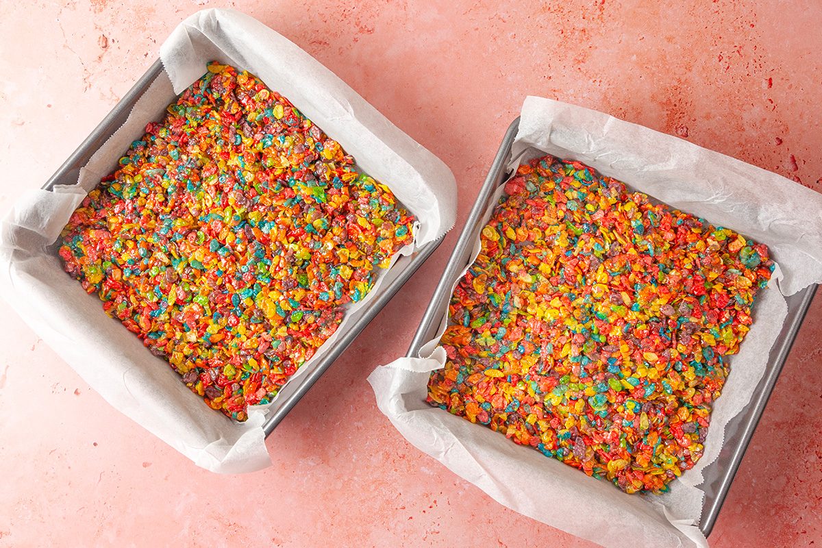Overhead shot of two square baking pans lined with parchment paper, each filled with colorful Fruity Pebbles cereal treats, on a pink surface