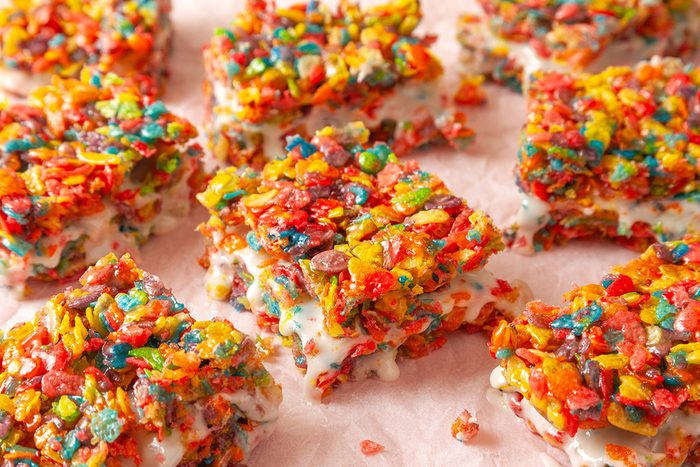 Overhead shot of Fruity Pebbles Milk 'n' Cereal Bars, cut into squares and set on pink parchment paper, with colorful cereal pieces and creamy white filling visible