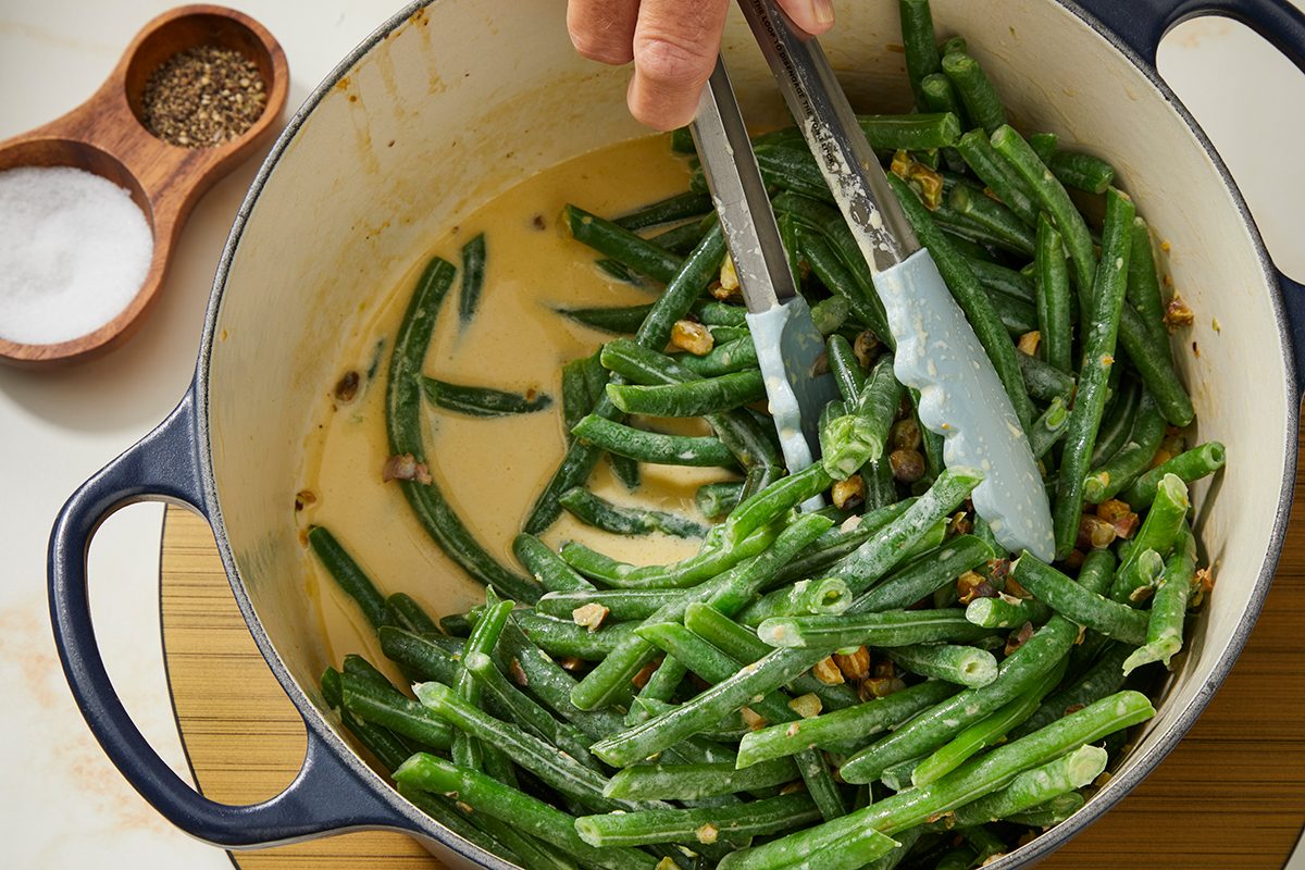 A hand uses tongs to mix cooked green beans and chopped garlic in a creamy sauce inside a blue Dutch oven. A small wooden dish with salt and pepper is nearby.