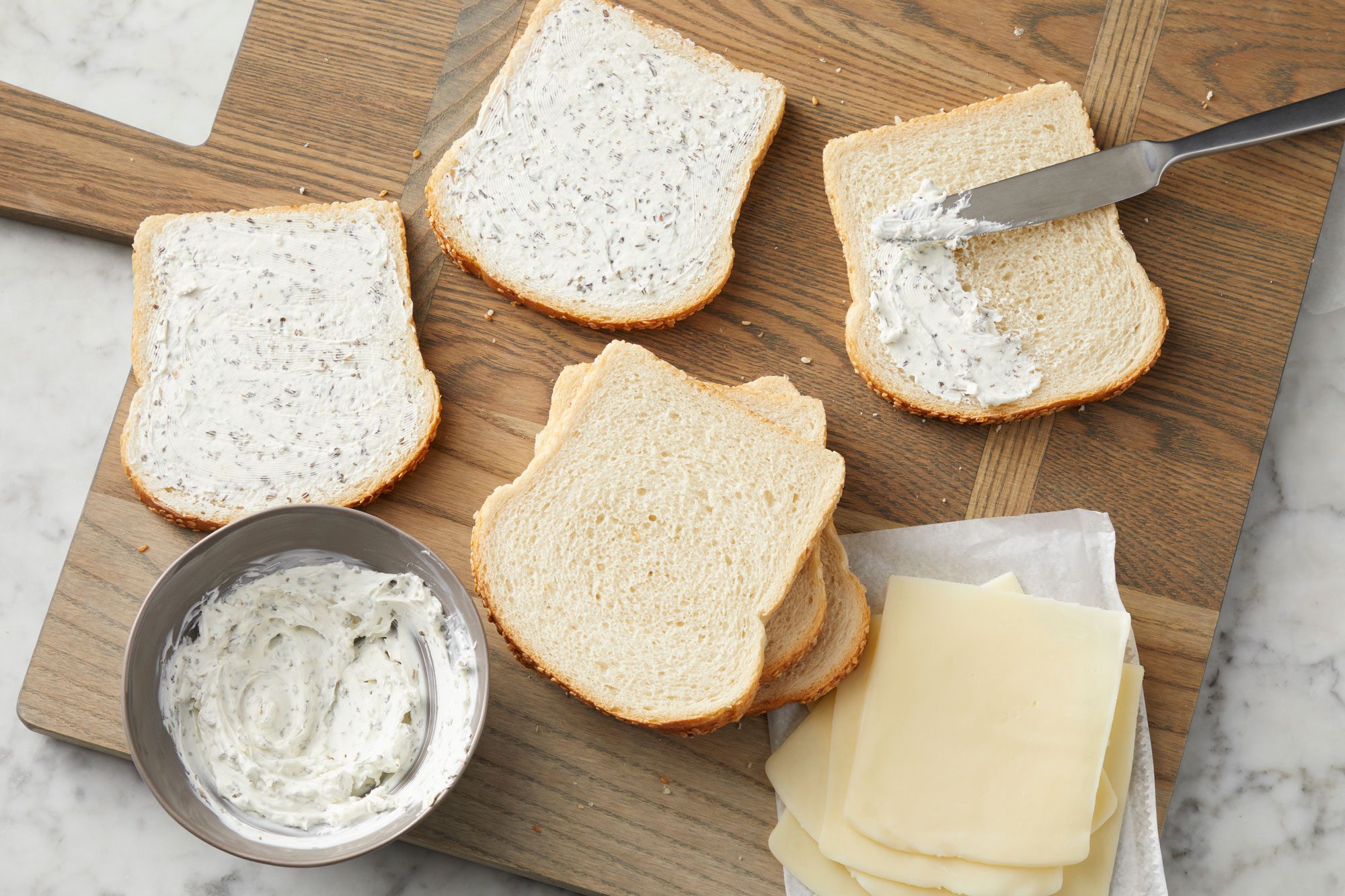 Overhead shot of a small bowl mix cream cheese and 1 teaspoon basil until blended; spread onto 6 bread slices on a wooden tray; Top with the mozzarella cheese and remaining bread; all set on a marble surface;