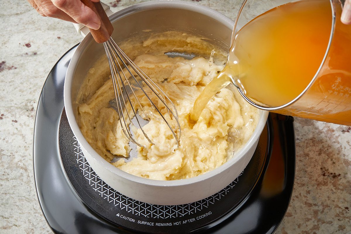 A person uses a whisk to mix a creamy roux in a saucepan while pouring broth from a measuring cup onto an electric stovetop.