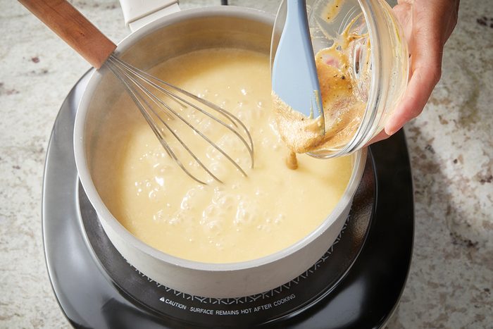 A hand pours a mixture from a small bowl into a pot of thick, creamy sauce on a stovetop. A whisk rests in the pot, and the scene is set on a marbled countertop.