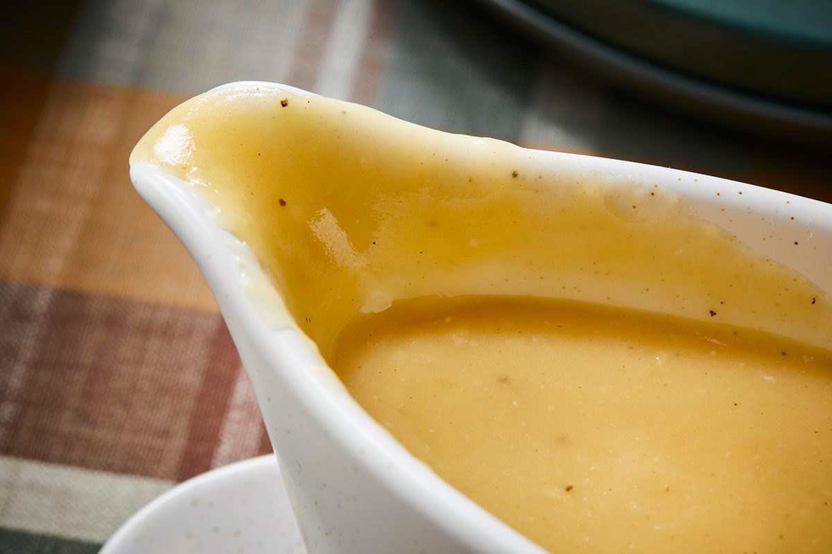 Close-up of a white gravy boat filled with smooth, golden-yellow gravy, showing a creamy texture with small black pepper specks, set on a plaid tablecloth background.