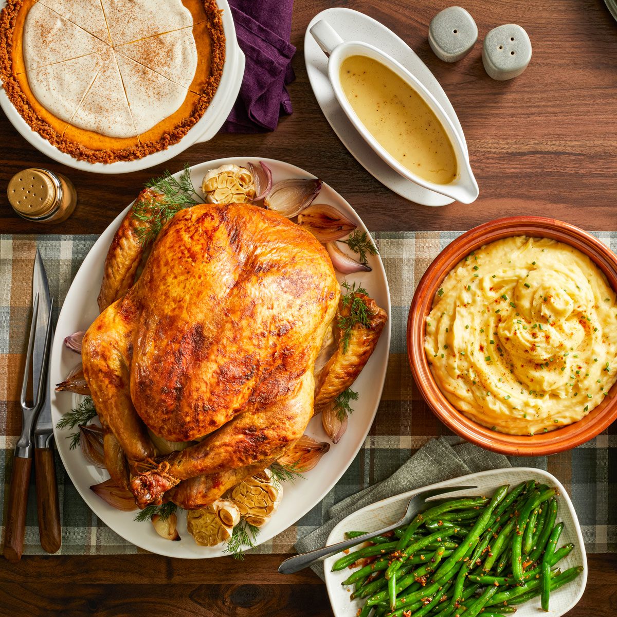 Overhead shot of a festive Thanksgiving meal arranged on a wooden table with a plaid runner, featuring golden Pickle-Brined Turkey, creamy Smoky Garlic Mashed Potatoes, Honey Garlic Green Beans, savory Sage Dressing, Aperol Spritz Cranberry Sauce, rich gravy, and Pumpkin Cheesecake Pie; Plates, utensils, and salt and pepper shakers complete the full holiday spread;