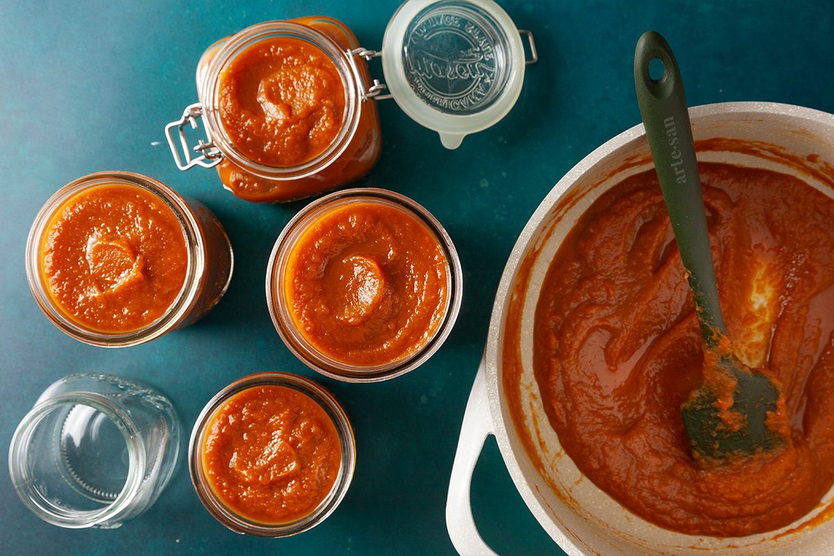 Several glass jars filled with smooth red tomato sauce are arranged on a blue surface next to a large white pot of sauce with a spatula inside. Some jars are open, and one is sealed with a hinged lid.