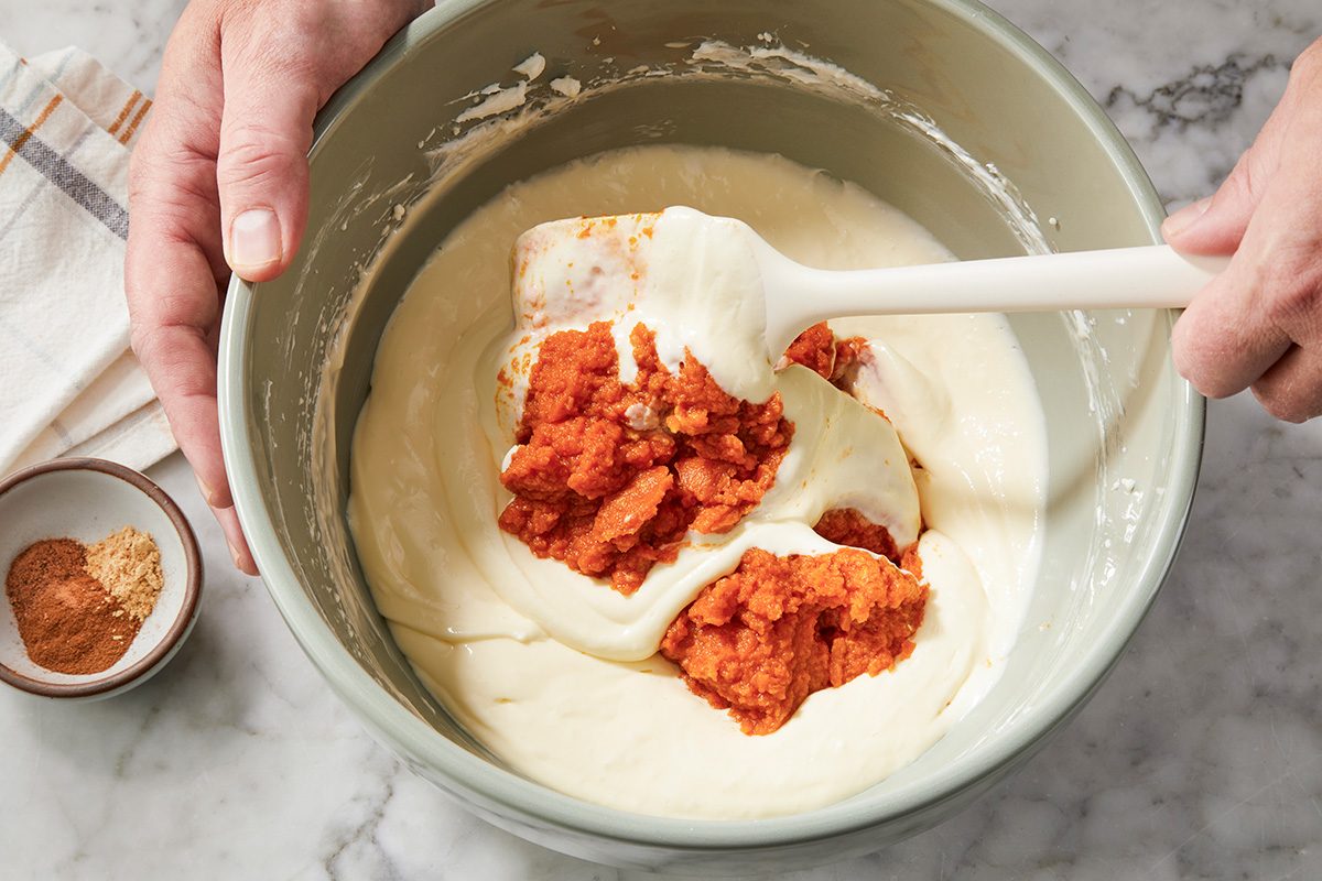A person stirs pumpkin puree into a creamy batter in a mixing bowl using a spatula. A small bowl of ground spices sits nearby on a marble countertop.
