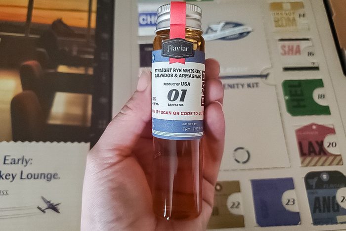 A hand holds a small bottle labeled “Barfar Straight Rye Whiskey, Series 01, Mission SEA” with a red seal. In the background, there are travel-themed cards and designs.