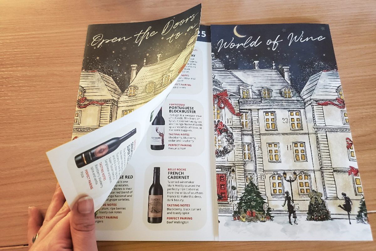 A hand turns the page of a festive wine catalog featuring illustrated holiday scenes with a snowy building, Christmas trees, and people outside, and descriptions of wine bottles inside.