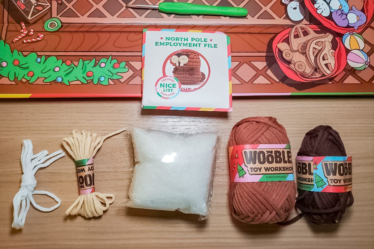 A craft kit is displayed on a table, including white string, yellow string, a stuffed white pouch, tan yarn, brown yarn, and a colorful instruction booklet labeled 