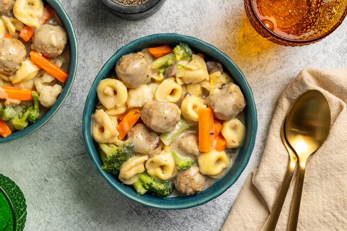 Overhead shot; bowl of tortellini pasta with meatballs; broccoli; carrots; cauliflower in creamy sauce; gold spoon and fork on beige napkin; glass of amber drink