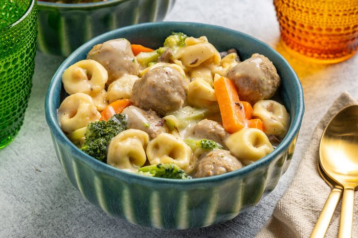 Overhead shot; blue bowl holds tortellini pasta; meatballs; broccoli; carrots in creamy sauce; gold spoon; napkin; textured glasses