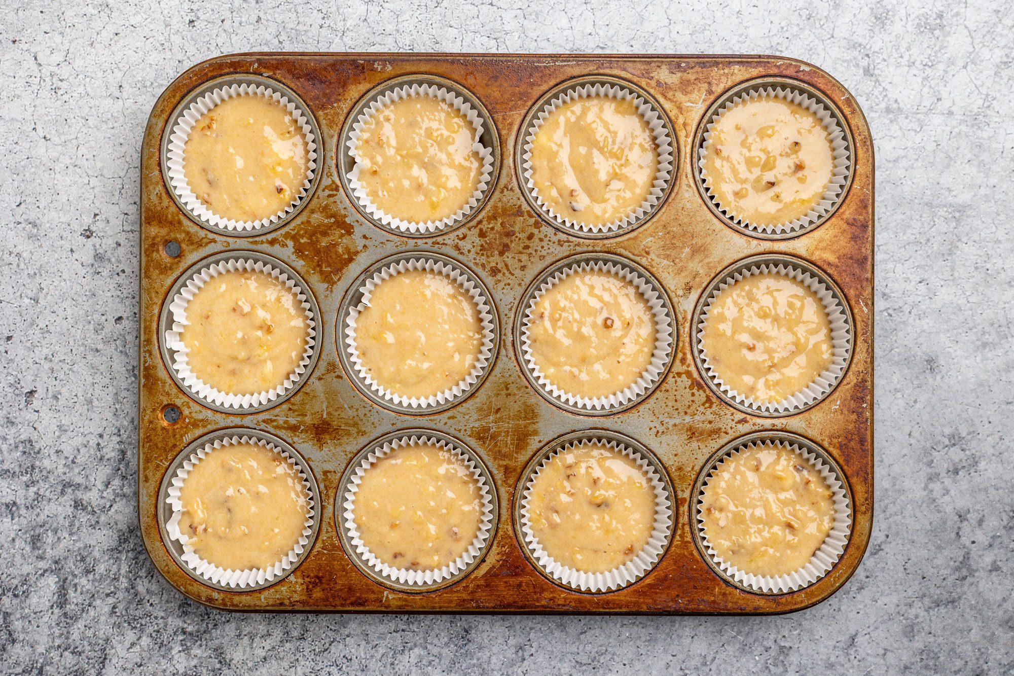 Overhead shot of a muffin tray with twelve paper liners, each filled with unbaked muffin batter, set on a gray textured surface; The tray shows a slightly worn, stained appearance