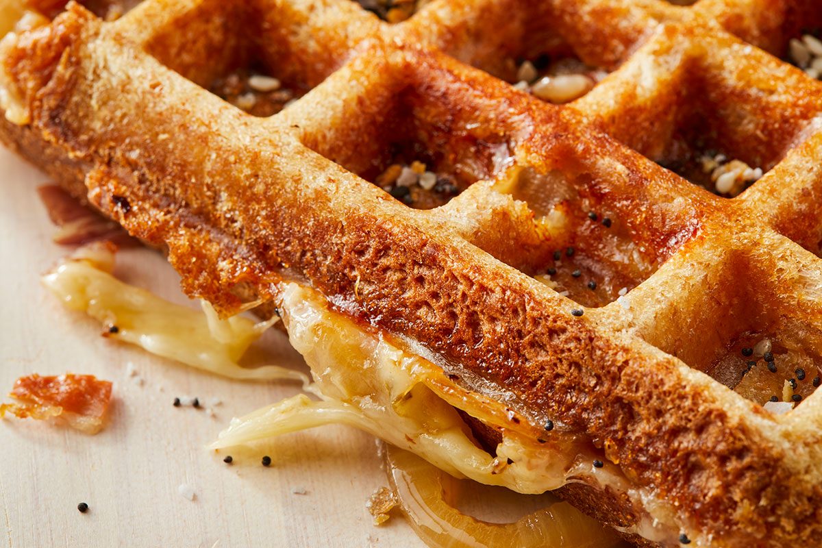 A close up shot of a crispy, golden brown waffle with melted cheese and seeds visible, showing a savory filling oozing out from between the waffle layers