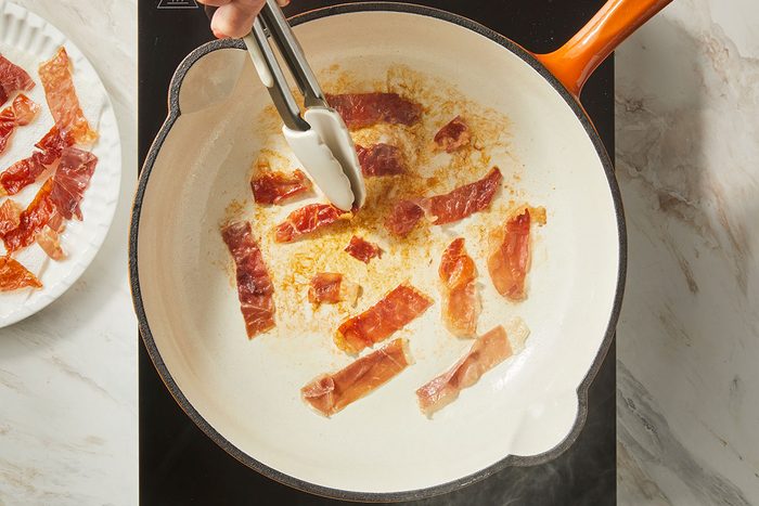 overhead shot of a hand uses tongs to cook strips of prosciutto in a white skillet on a stovetop, with more prosciutto pieces on a plate nearby
