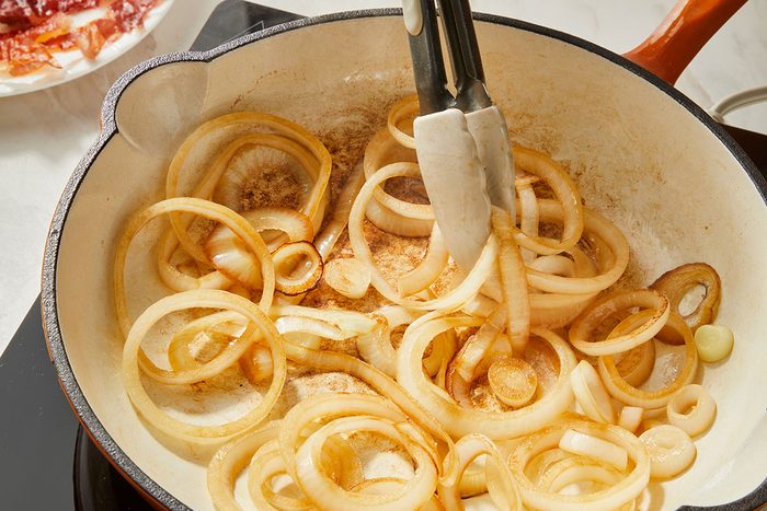 3/4th shot of a pair of tongs stirs sliced onions in a white cast iron skillet on a stovetop; cooked bacon is visible on a plate in the background