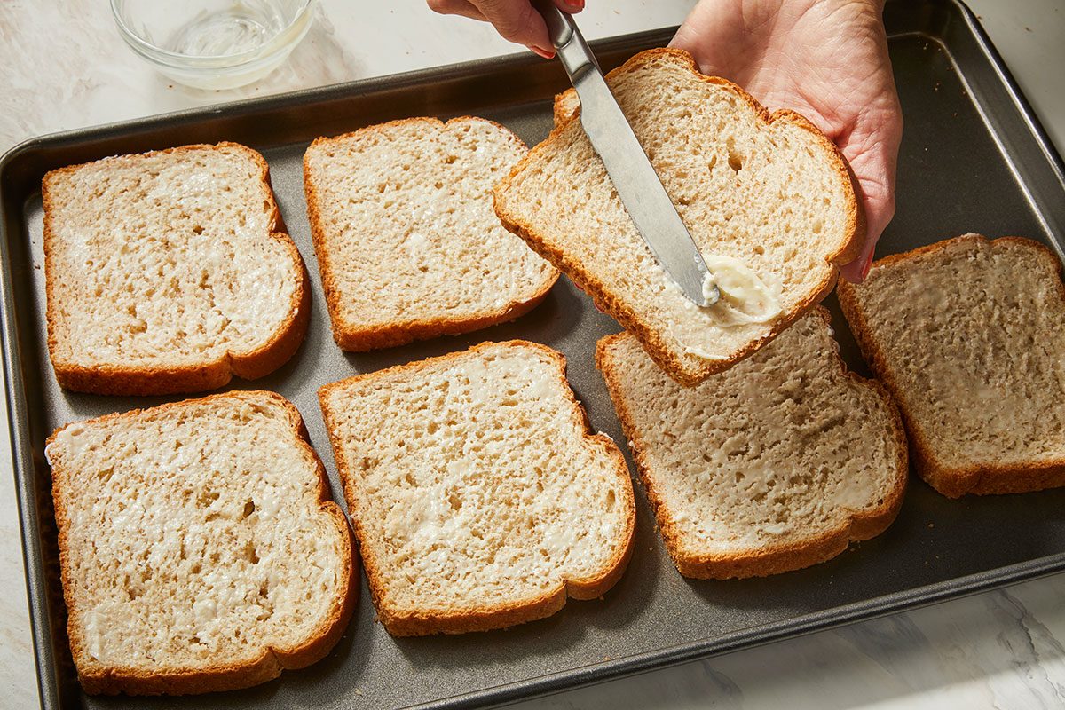 3/4th shot of a hand spreads butter on a slice of bread with a knife over a baking tray holding several other buttered bread slices