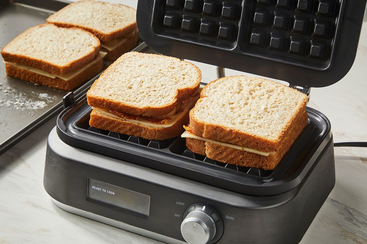 3/4th shot of wo sandwiches on wheat bread are being grilled in an open panini press, while two more sandwiches sit on a baking tray nearby; The scene is set on a light countertop